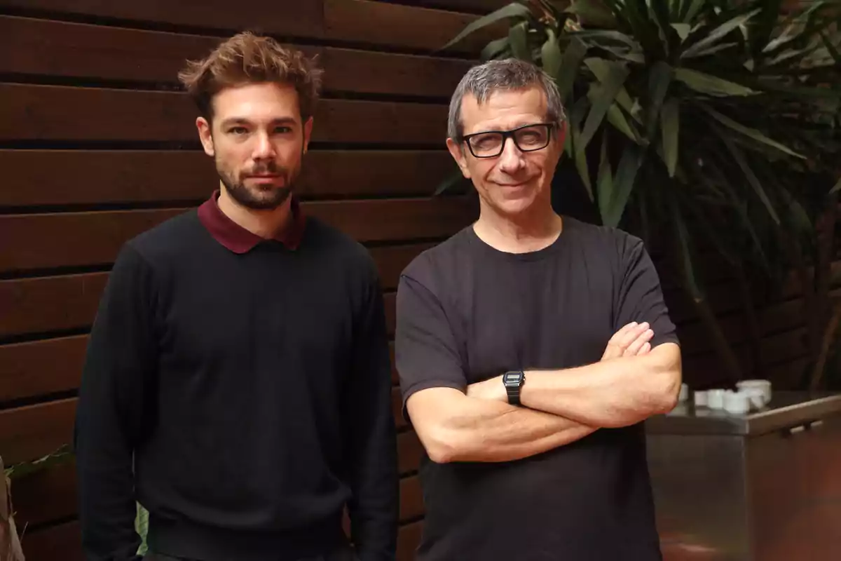 Dos hombres posan juntos frente a una pared de madera y plantas.