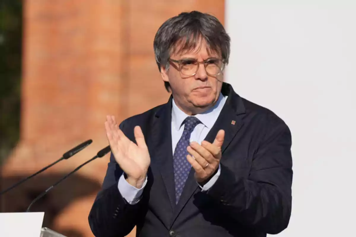 Un home amb ulleres i vestit fosc aplaudint mentre està dret davant d'un micròfon. Un home amb ulleres i vestit fosc aplaudint mentre està dret davant d'un micròfon.