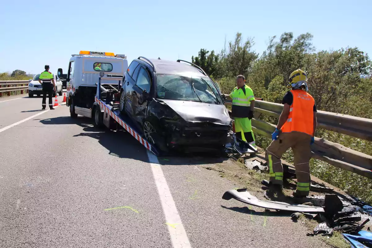 Un coche dañado está siendo remolcado por una grúa en una carretera mientras dos personas con chalecos de seguridad observan la escena.