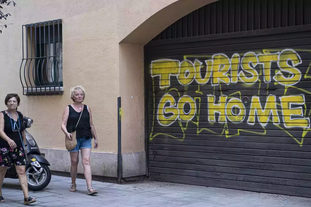 Dues dones caminant per un carrer al costat d'una porta de garatge amb un grafit que diu “TOURISTS GO HOME”.