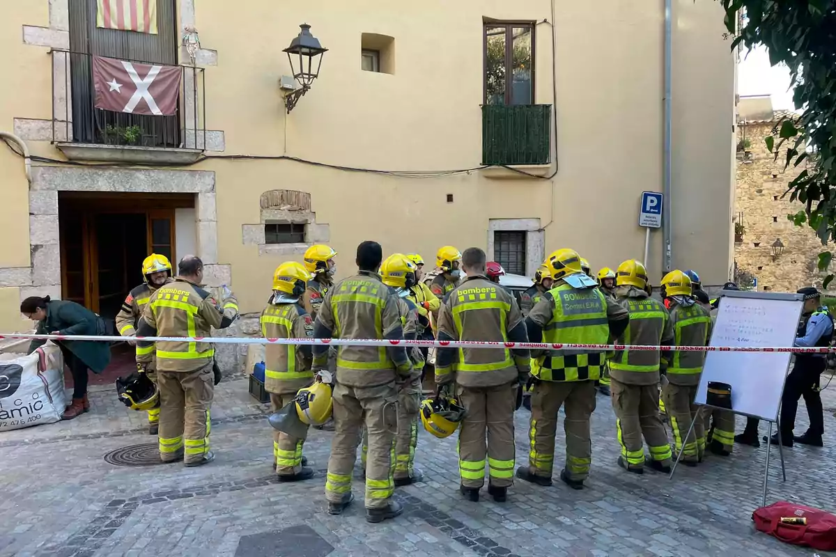 Un grup de bombers amb cascos grocs es troben reunits davant d'un edifici, mentre una persona s'inclina a prop d'una bossa gran i un tauler blanc està col·locat de banda.