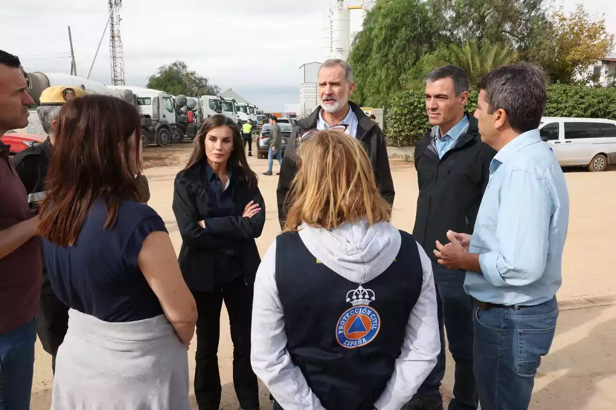 Un grupo de personas conversando al aire libre en un área con vehículos y árboles al fondo.