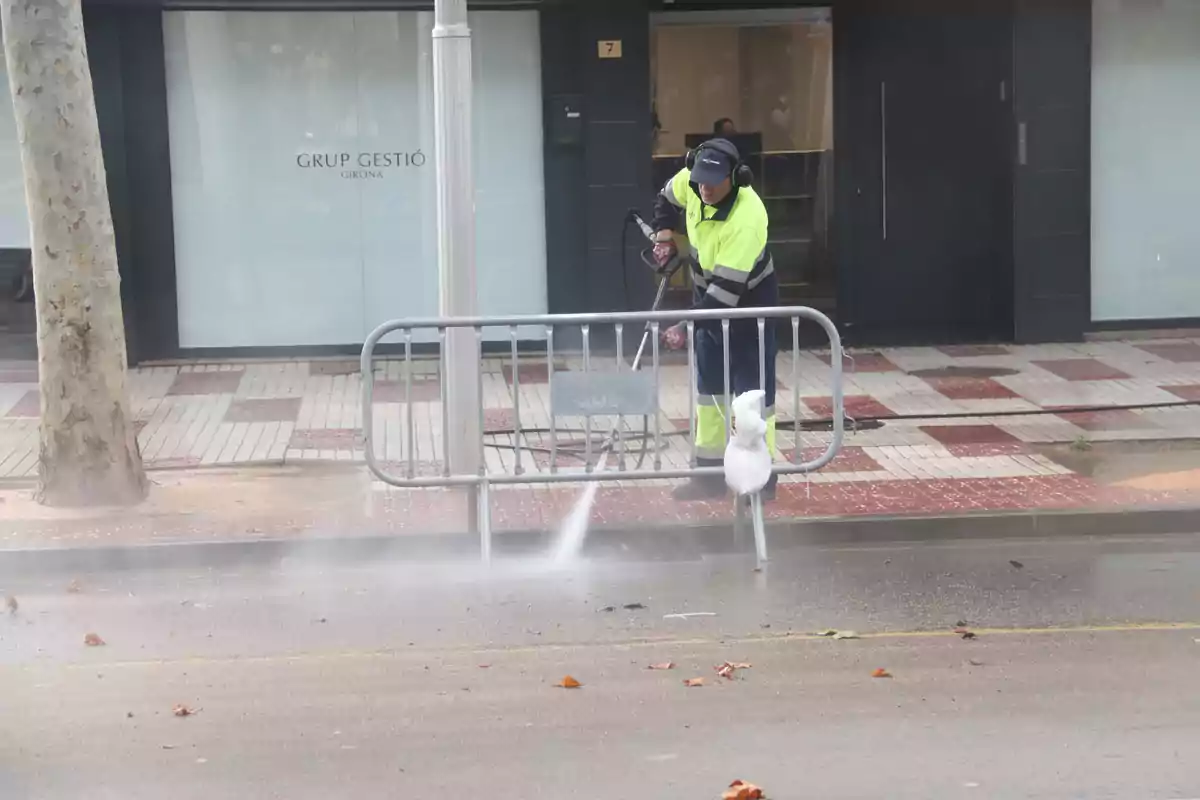 Un treballador de neteja amb uniforme d'alta visibilitat utilitza una hidrolavadora per netejar el carrer davant d'un edifici amb el rètol "Grup Gestió Girona".