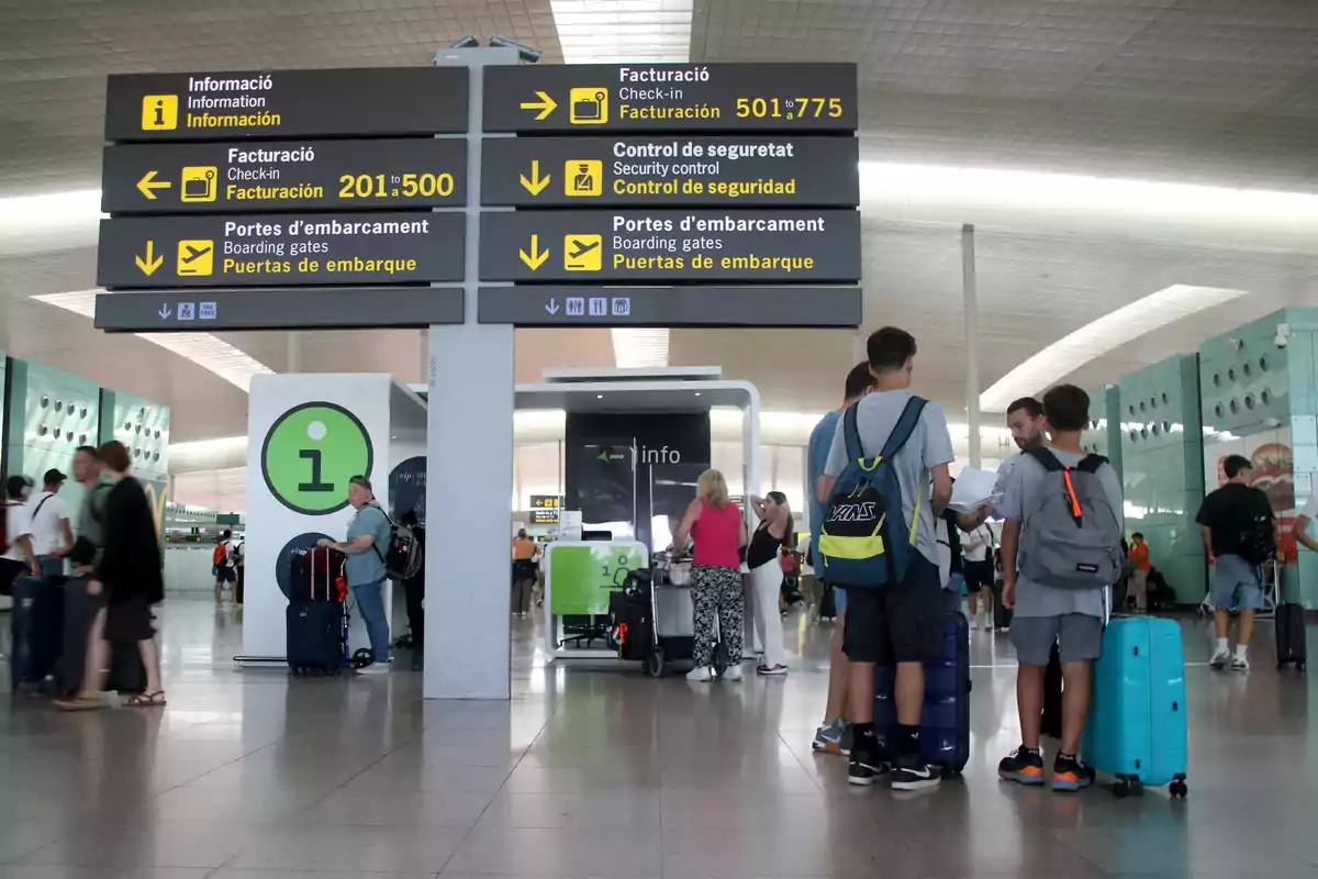 Persones a un aeroport observen senyals d'informació, facturació, control de seguretat i portes d'embarcament, amb maletes i motxilles mentre esperen.