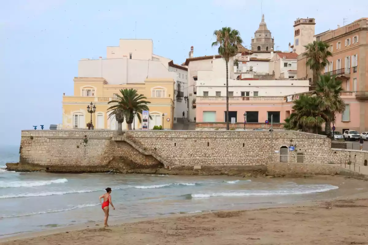 Una mujer en traje de baño rojo camina por la orilla de una playa con edificios históricos y palmeras al fondo.