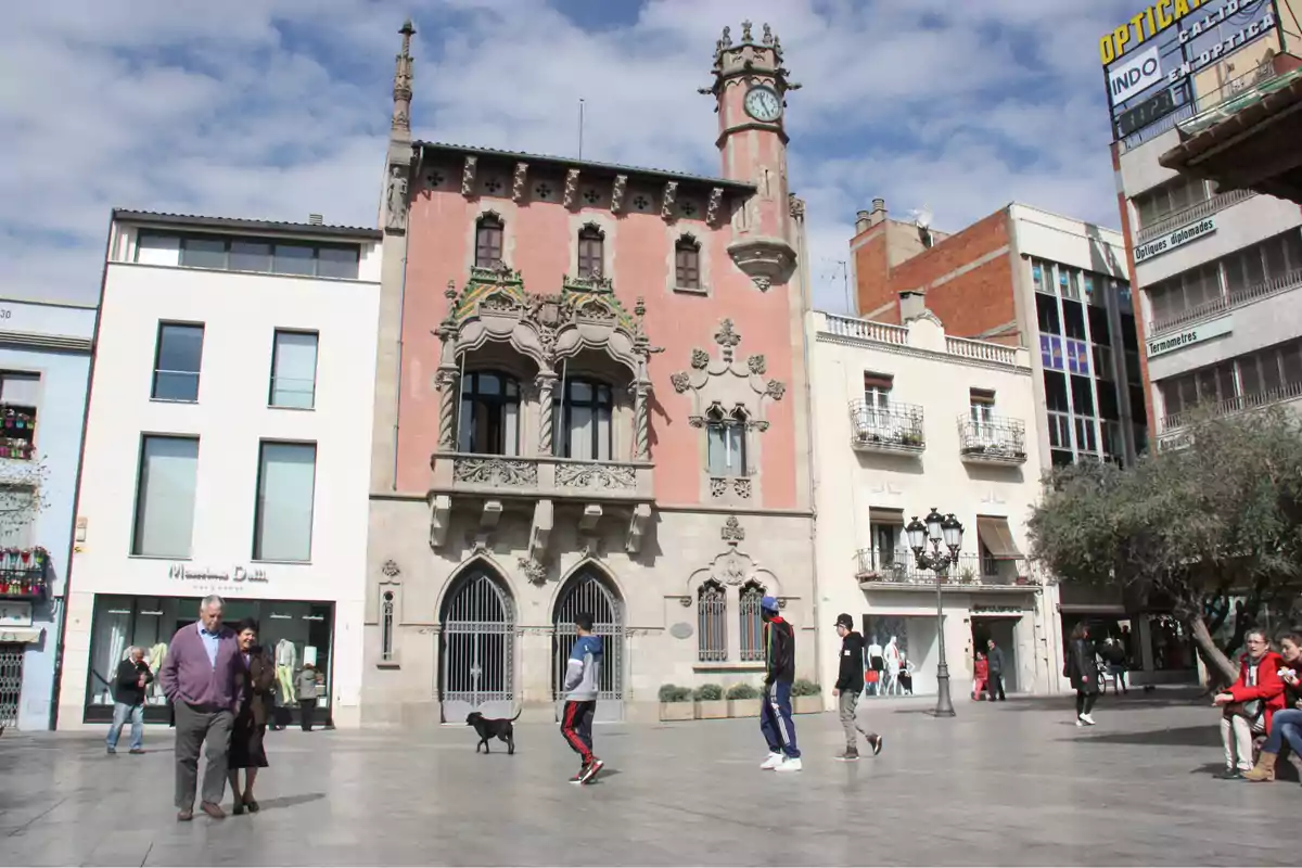 Persones caminant i assegudes a una plaça amb edificis històrics i moderns al voltant en un dia assolellat.