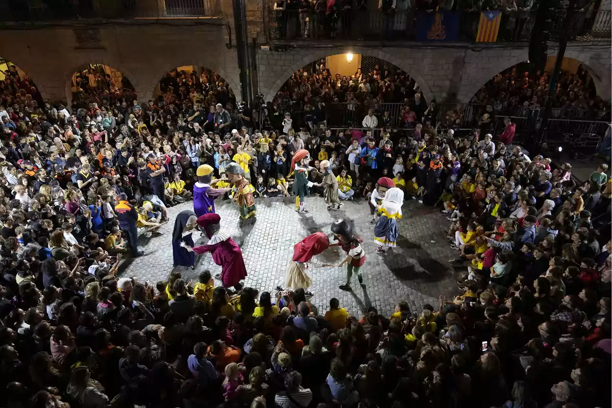 Una multitud observa un espectacle de persones disfressades amb capgrossos en una plaça il·luminada a la nit.