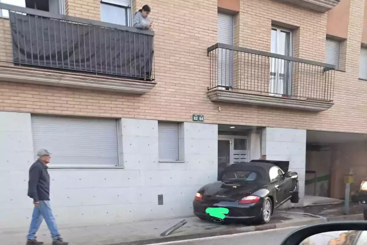 Un cotxe esportiu negre està parcialment pujat a la vorera davant d'un edifici de maó mentre un home camina per la vorera i una altra persona observa des d'un balcó.