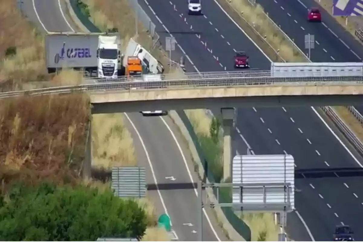 Un camión accidentado en la salida de una autopista junto a un puente y varios vehículos circulando por la carretera principal