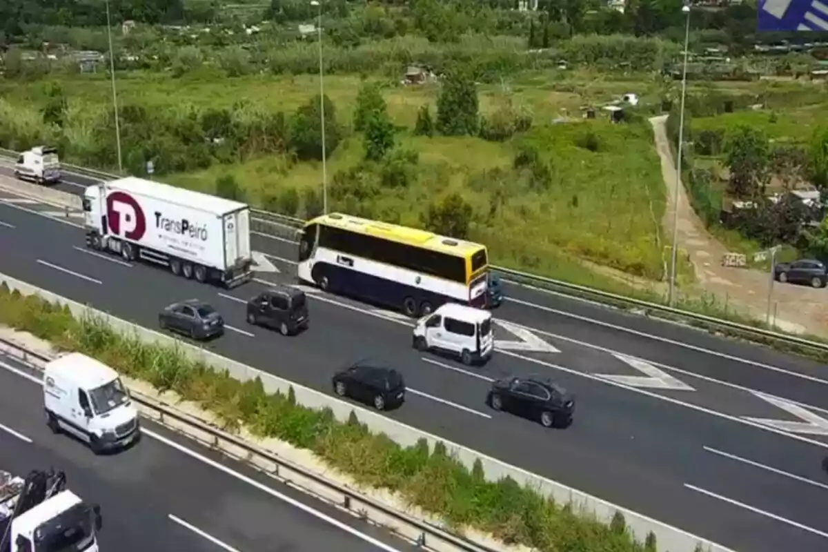 Un autobús groc i blanc es troba aturat a la vorera d'una autopista mentre diversos vehicles circulen pels carrils adjacents envoltats d'un paisatge verd.