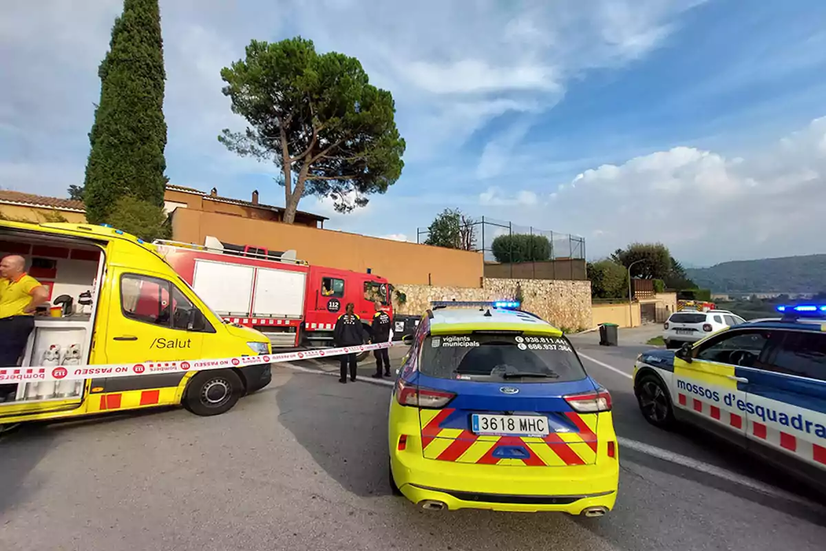 Vehicles d'emergència i personal en un carrer acordonat davant d'una casa amb un arbre gran i cel parcialment ennuvolat.