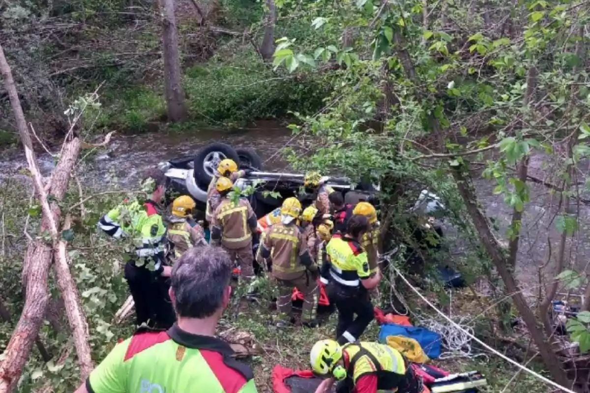 Un grup de rescatadors treballa a l'escena d'un accident on un vehicle ha bolcat a prop d'un riu en una àrea boscosa.