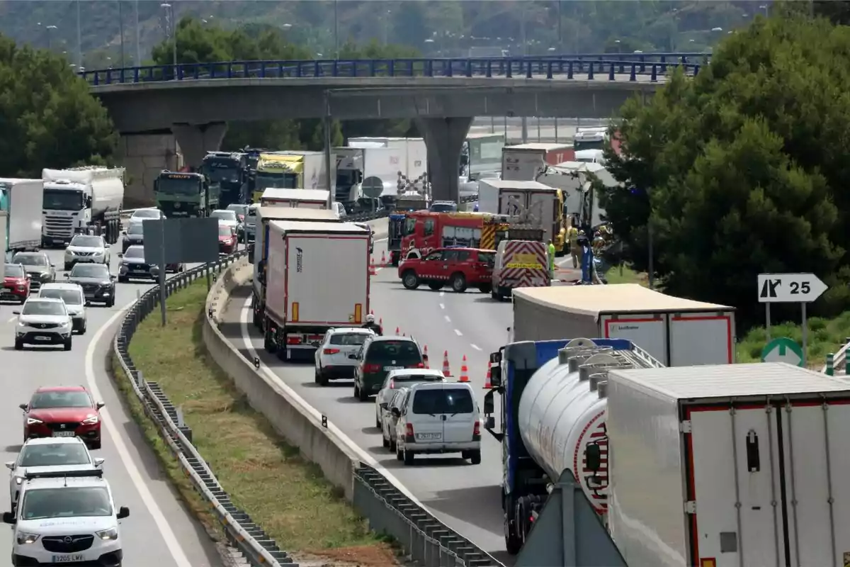 Tráfico detenido en una autopista debido a un accidente, con varios camiones y coches en la carretera y vehículos de emergencia en la escena.