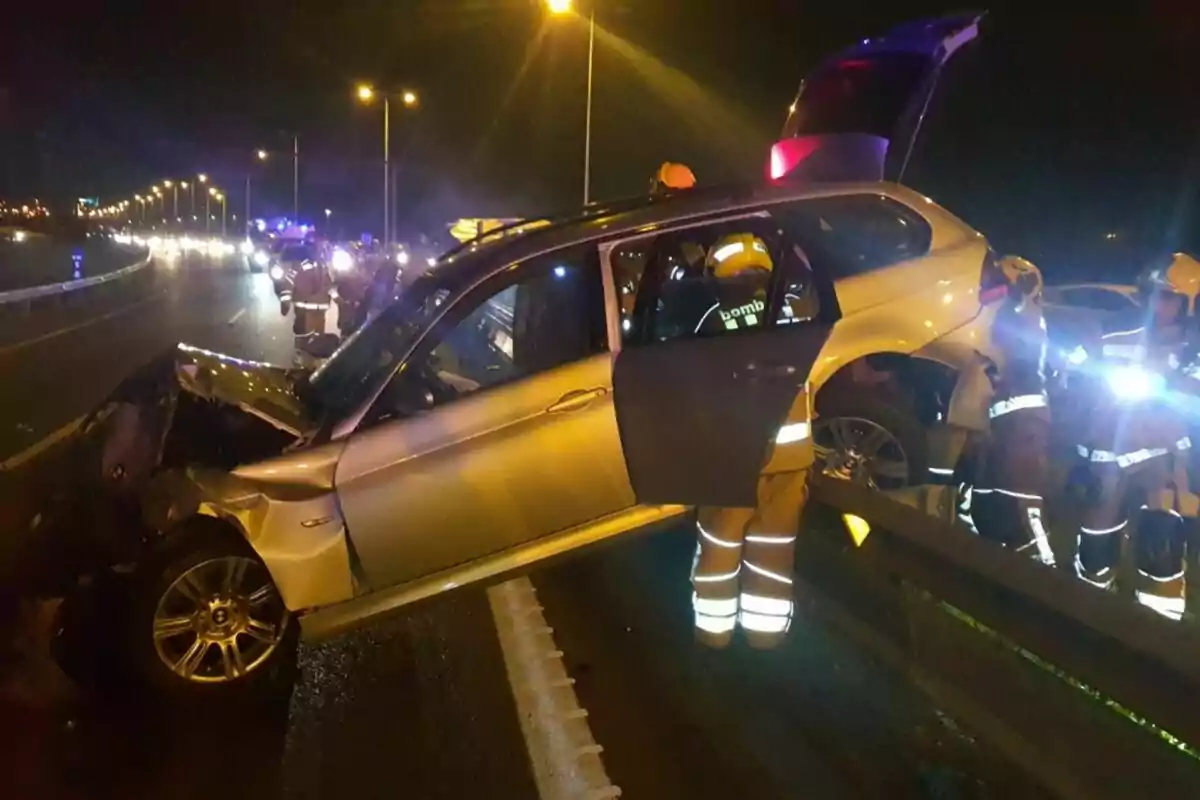 Un coche accidentado sobre una barrera en una carretera de noche con bomberos alrededor iluminando la escena.