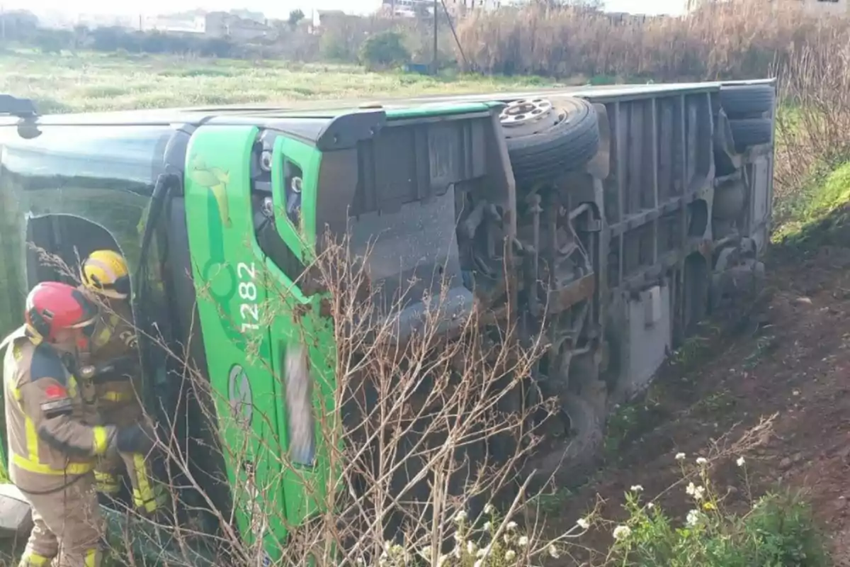 Un autobús verde volcado de lado en un campo con dos bomberos trabajando en la escena.