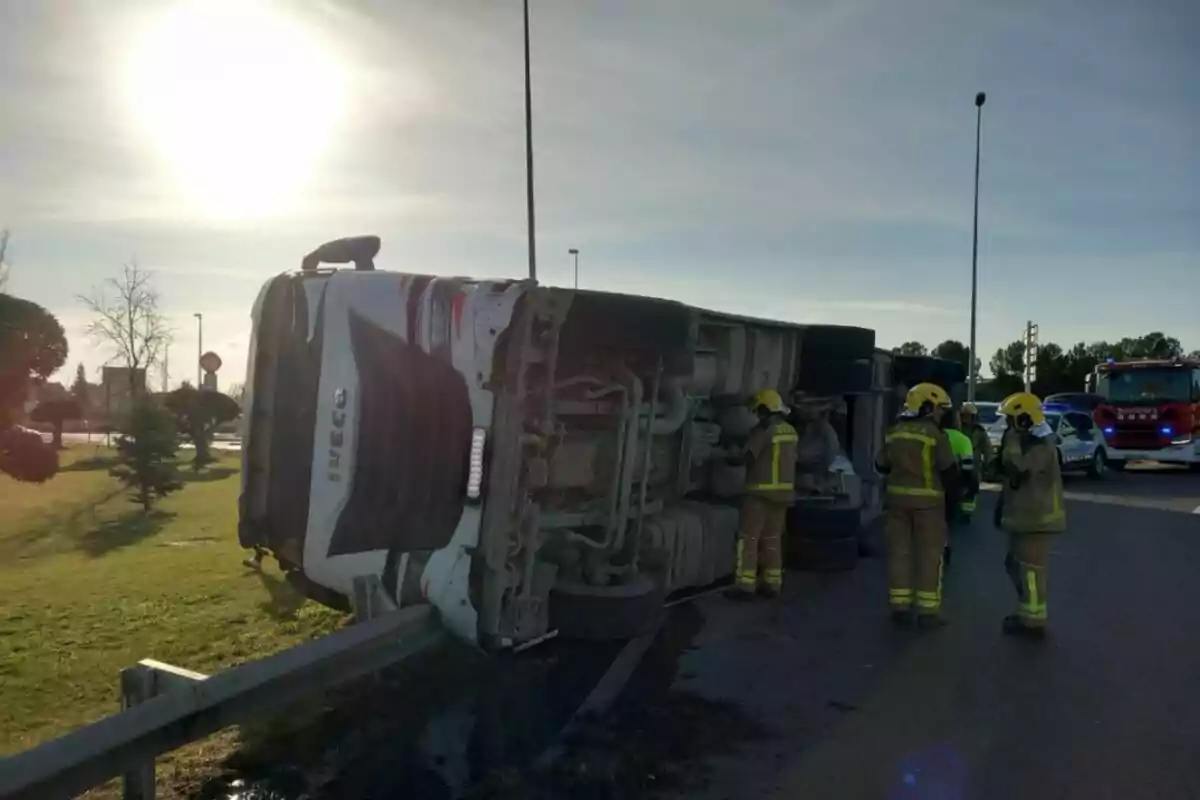 Un camión volcado en la carretera con bomberos trabajando en el lugar y un camión de bomberos al fondo.