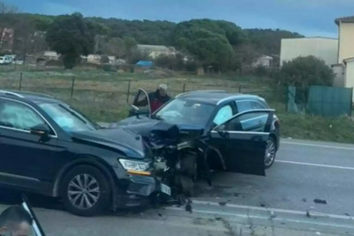 Dos cotxes negres xocats de front en una carretera amb una persona dreta al costat d'un dels vehicles i un paisatge rural al fons.