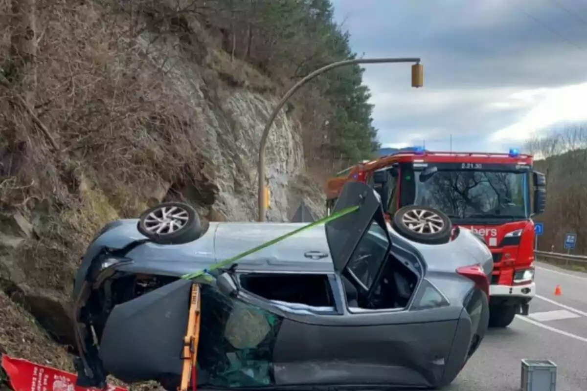 Un cotxe bolcat a la carretera amb un camió de bombers al fons.