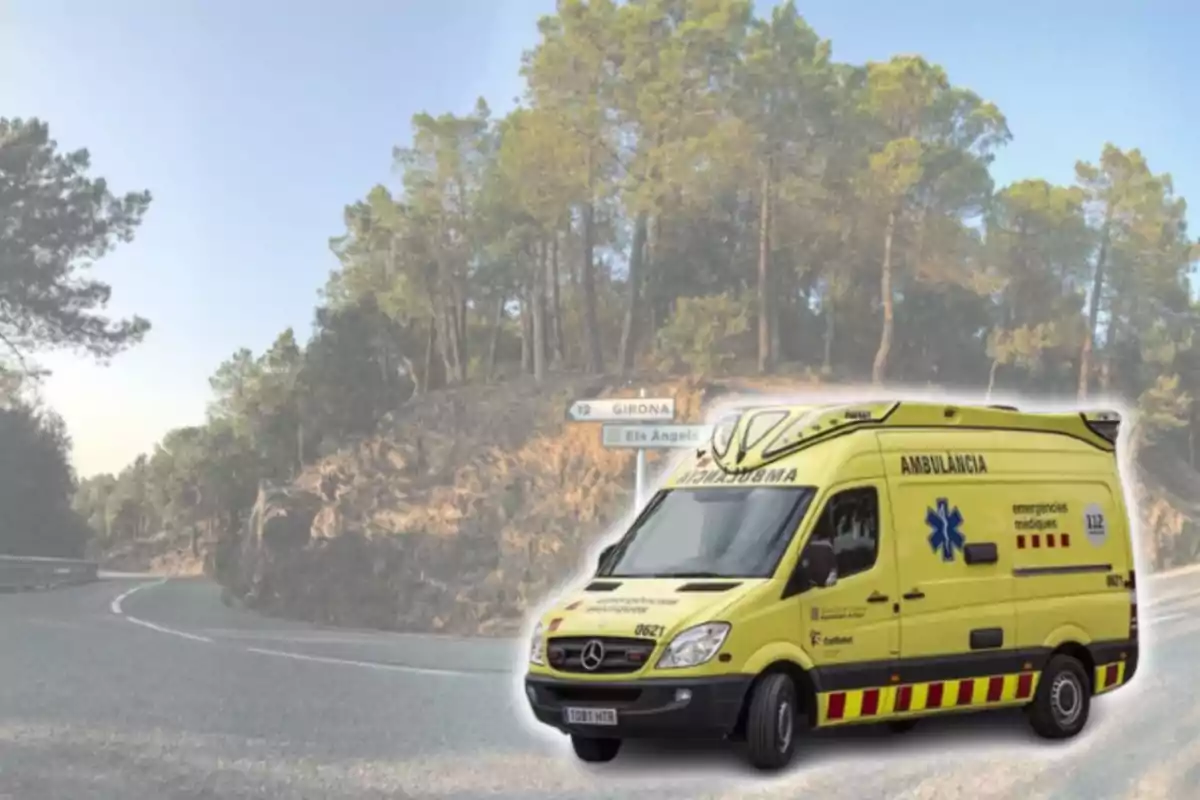 Una ambulancia amarilla está estacionada en una carretera rodeada de árboles con un cartel que indica direcciones hacia Girona y Els Àngels.