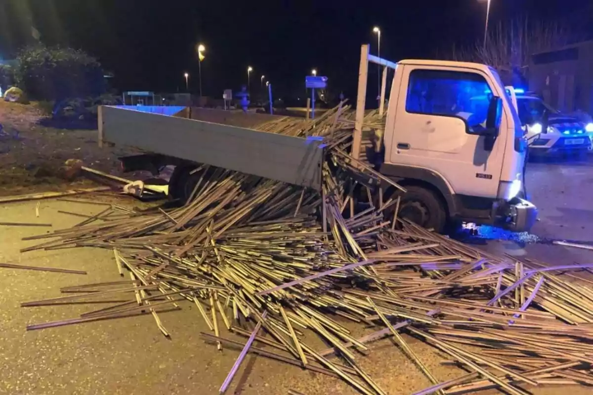 Un camión volcado en la carretera con barras de metal esparcidas por el suelo durante la noche.