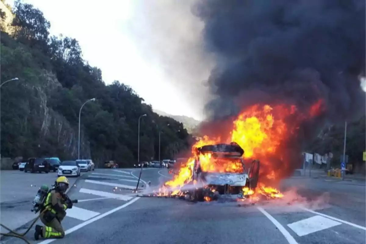 Un bomber combat un incendi en un vehicle en flames en una carretera, amb fum negre elevant-se al cel i diversos cotxes estacionats al fons.