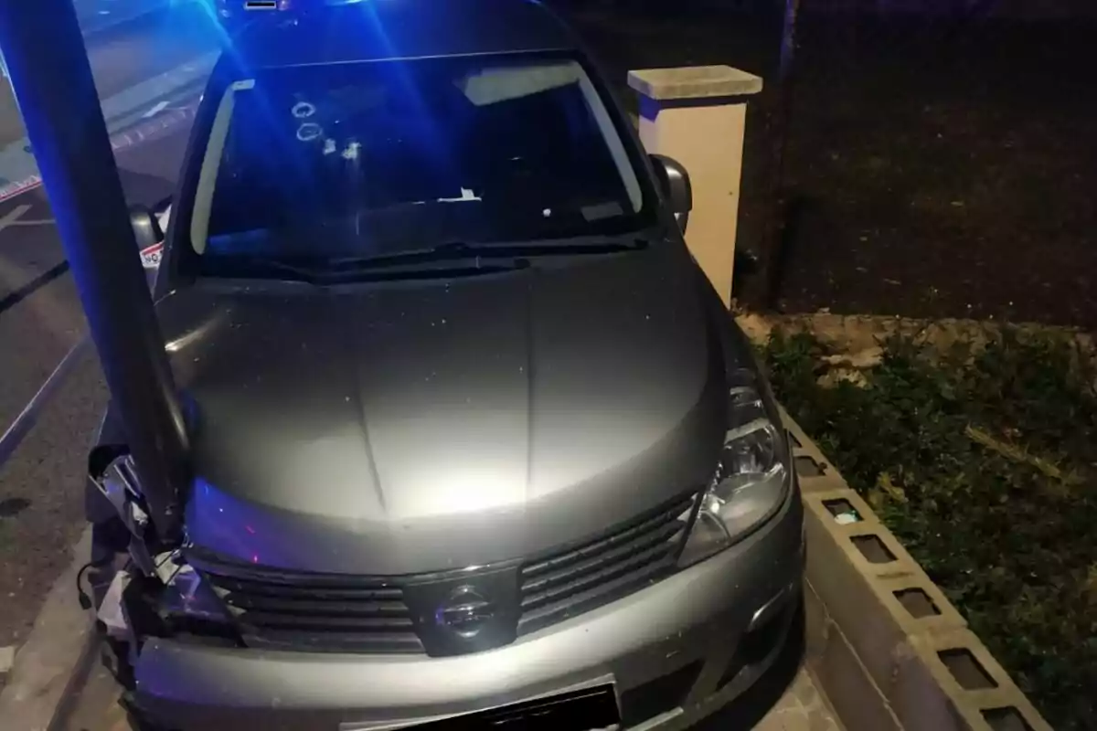 Un coche gris con el frente dañado está detenido contra un poste en una calle iluminada por luces azules.