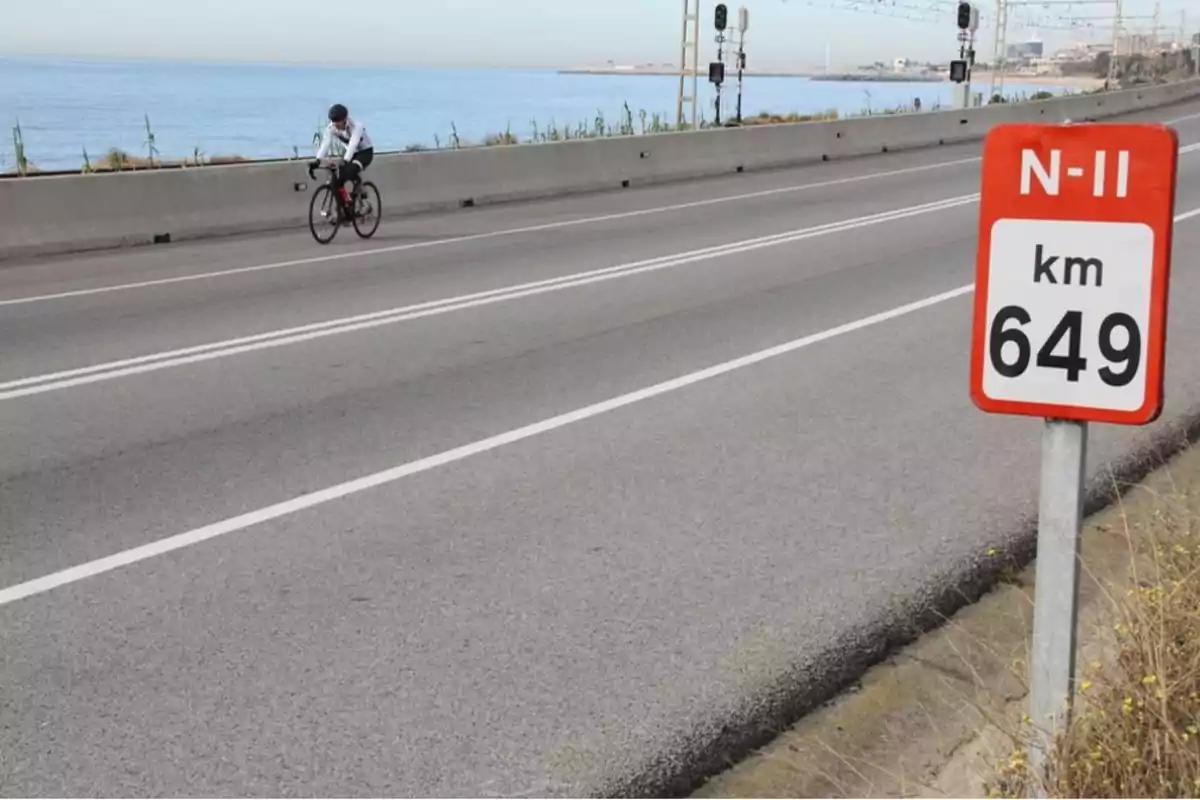 Un ciclista pedala per una carretera al costat del mar amb un senyal que indica el quilòmetre 649 de la N-II