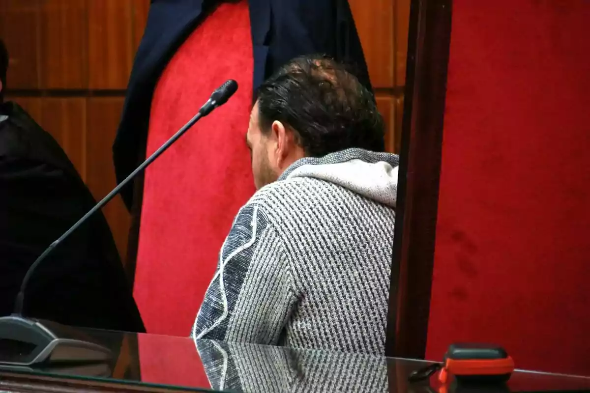 Un hombre sentado de espaldas en una sala con sillas rojas y un micrófono frente a él.