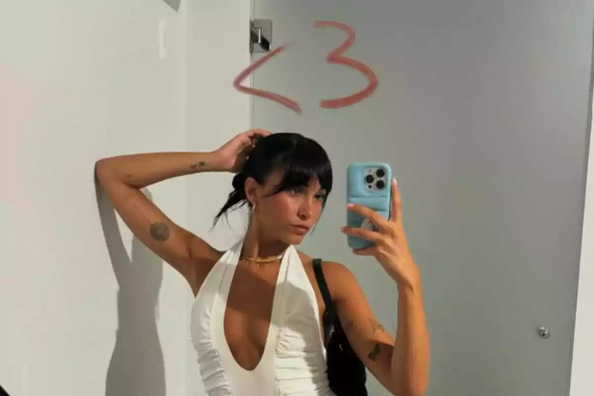 Mujer con vestido blanco tomándose una selfie frente a un espejo con un dibujo de corazón en la pared.