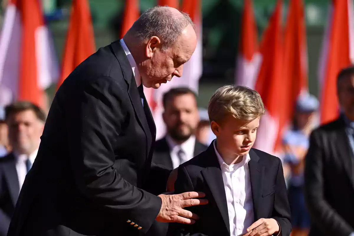 Albert of Monaco and Jacques, both dressed in dark suits, are standing at an outdoor formal event with red and white flags in the background. Albert of Monaco and Jacques, both dressed in dark suits, are standing at an outdoor formal event with red and white flags in the background.