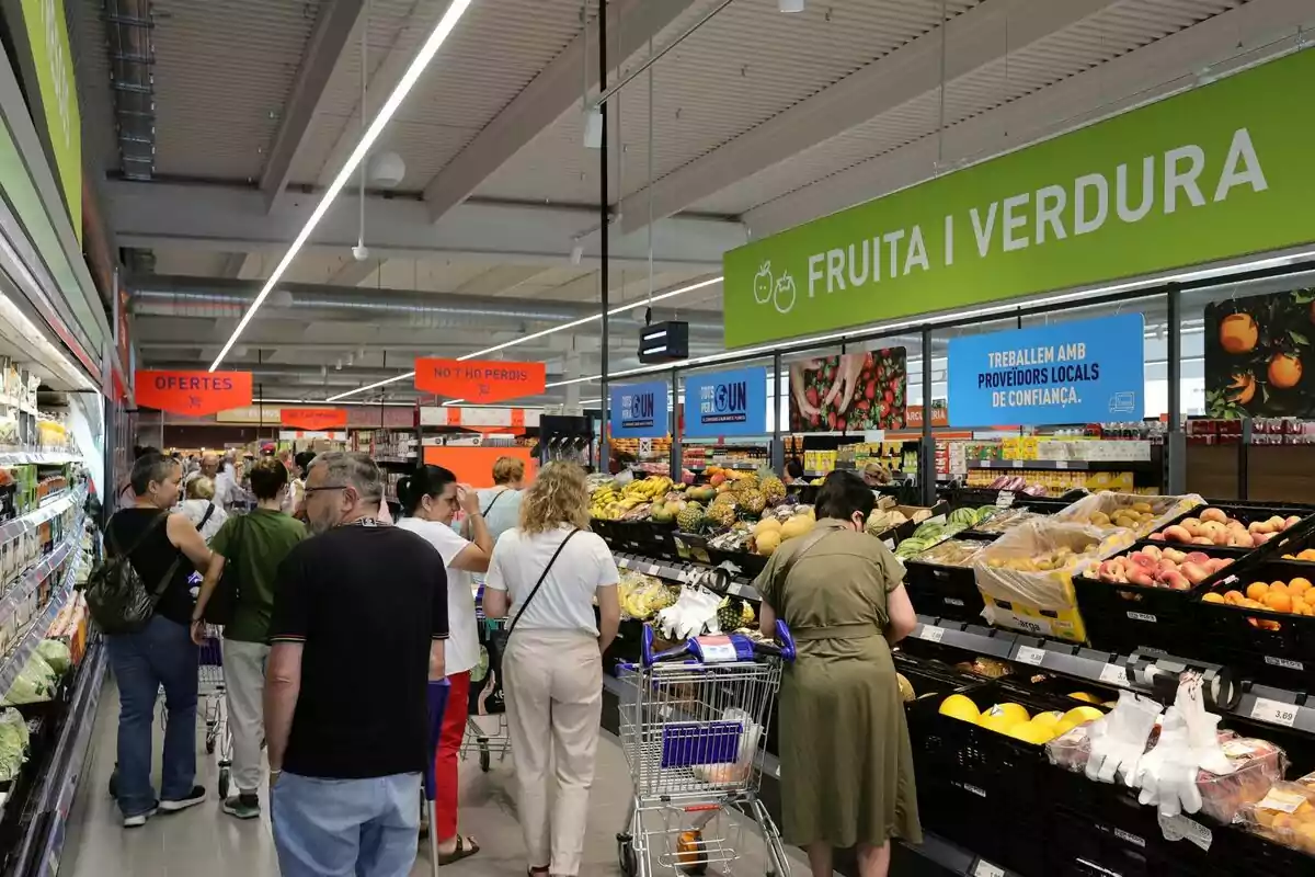 Personas comprando en la sección de frutas y verduras de un supermercado, con letreros en catalán que indican ofertas y productos locales.