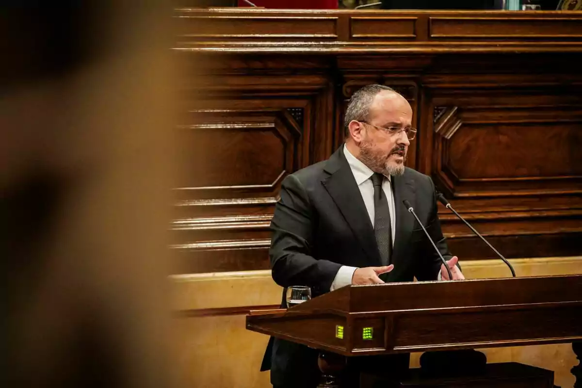 Un home amb vestit fosc parla en un faristol de fusta en un entorn formal. Un home amb vestit fosc parla en un faristol de fusta en un entorn formal.