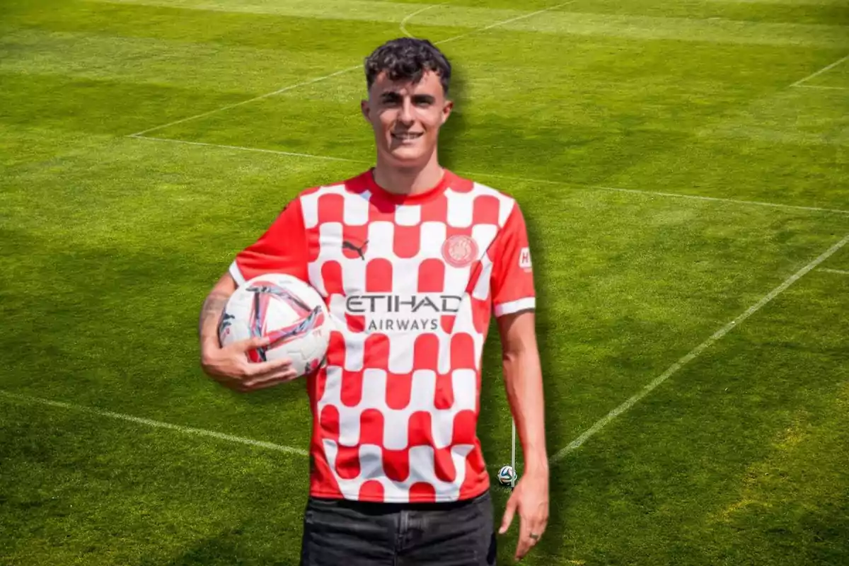 Un jugador de fútbol con una camiseta roja y blanca a cuadros sostiene un balón en un campo de fútbol.