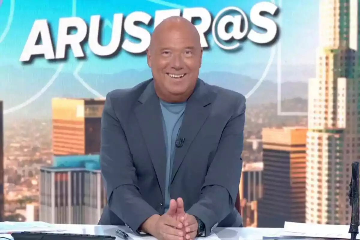 Un presentador sonriente en un set de televisión con un fondo urbano y el logo de un programa detrás.