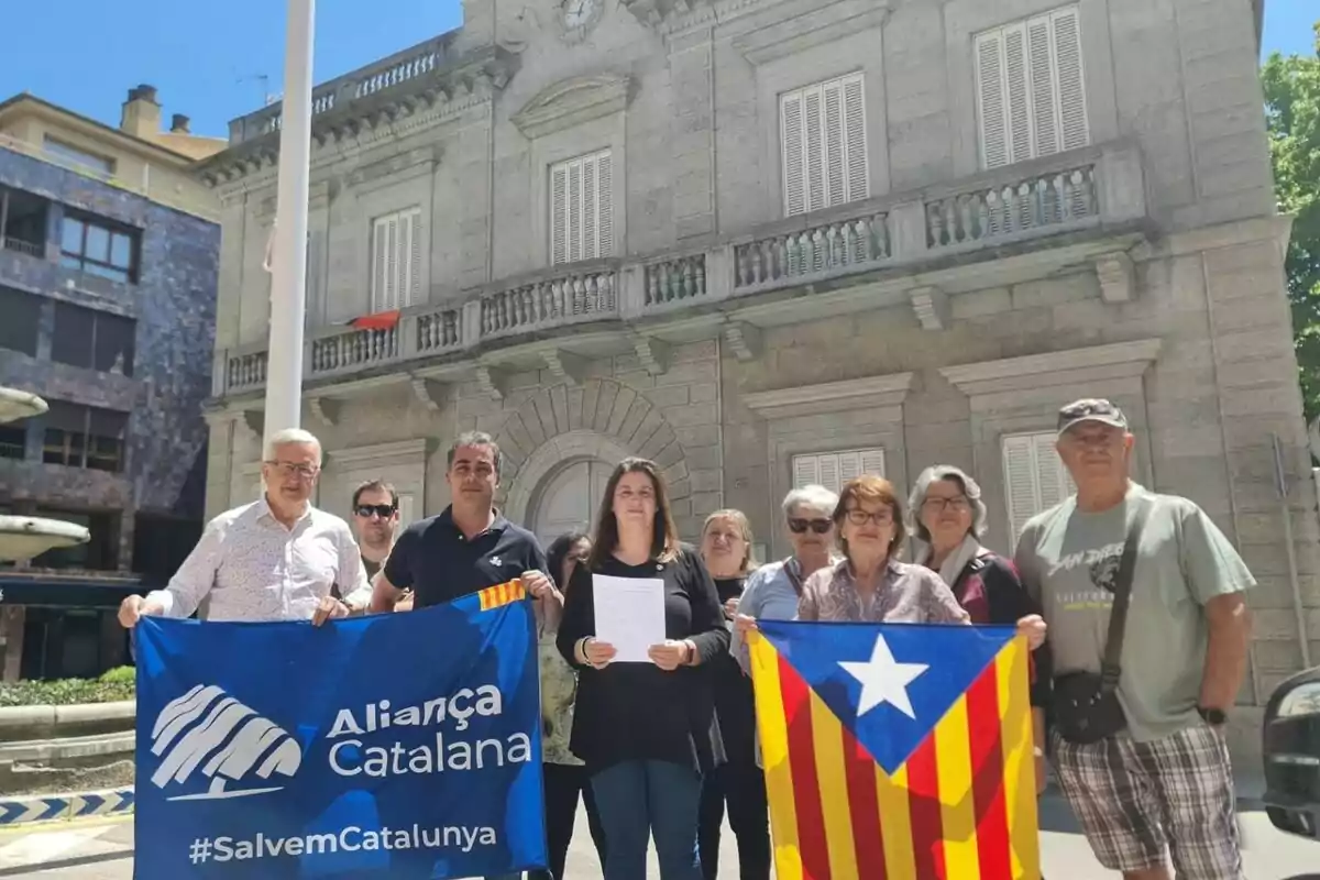 Un grup de persones posa davant d’un edifici antic sostenint una bandera blava d’Aliança Catalana i una estelada catalana.