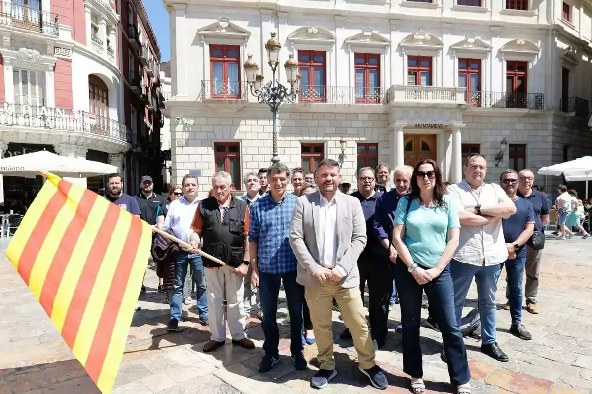 Un grup de persones posant en una plaça davant d’un edifici amb finestres vermelles i una bandera groga amb franges vermelles en primer pla Un grup de persones posant en una plaça davant d’un edifici amb finestres vermelles i una bandera groga amb franges vermelles en primer pla