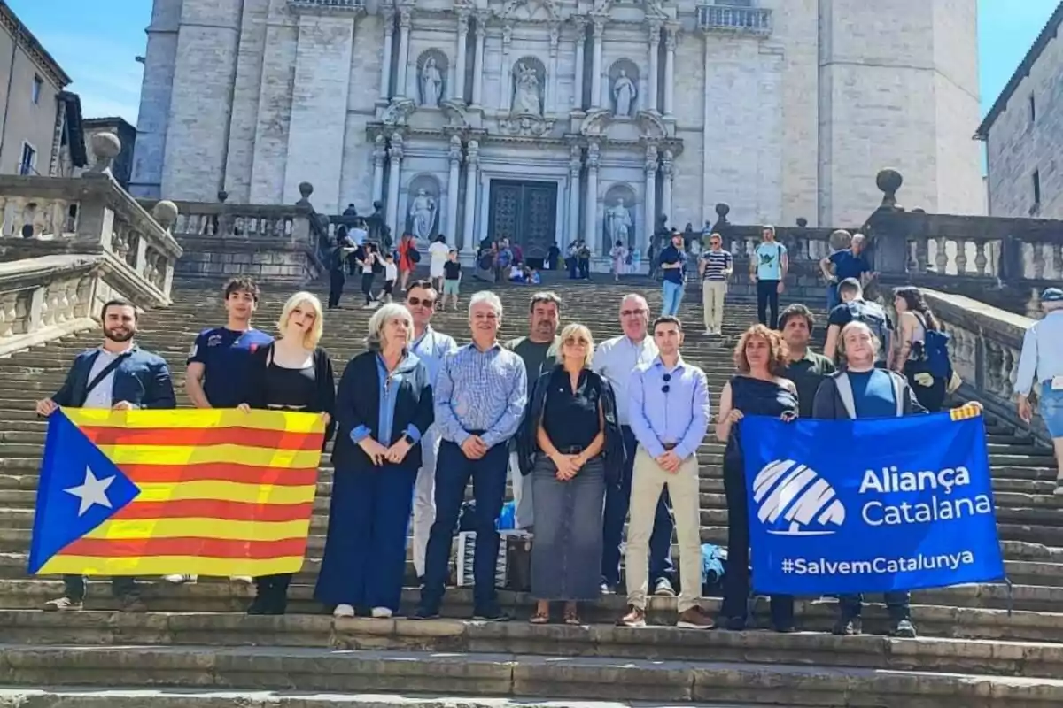 Un grup de persones posant a les escales d’una església, sostenint una bandera estelada i una pancarta blava d’Aliança Catalana. Un grup de persones posant a les escales d’una església, sostenint una bandera estelada i una pancarta blava d’Aliança Catalana.