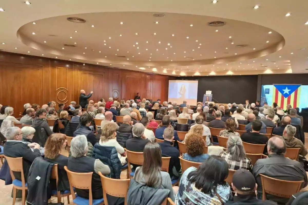 Una sala de conferències plena de persones assegudes, amb una pantalla al davant mostrant una presentació i una bandera amb una estrella blanca sobre un fons blau i franges grogues i vermelles a la dreta.