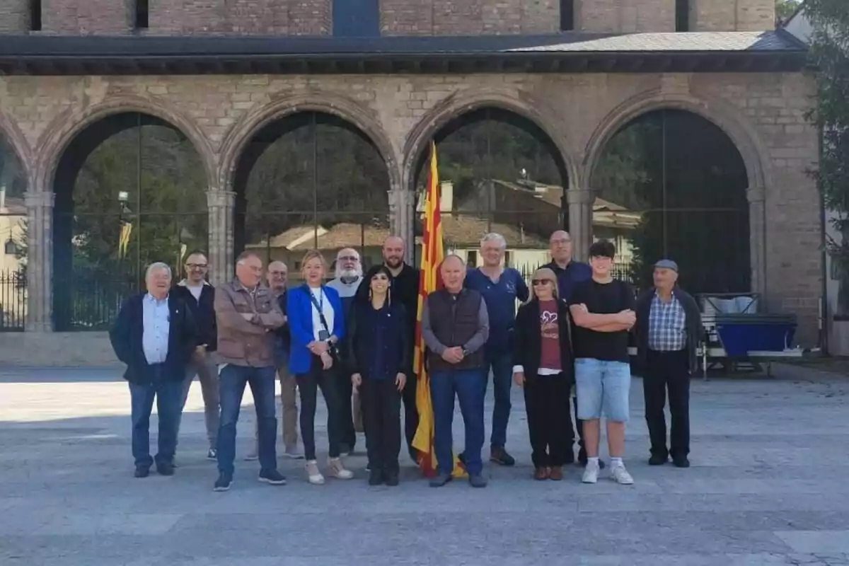 Un grup de persones posant davant d’un edifici de pedra amb arcs i una bandera catalana al centre Un grup de persones posant davant d’un edifici de pedra amb arcs i una bandera catalana al centre