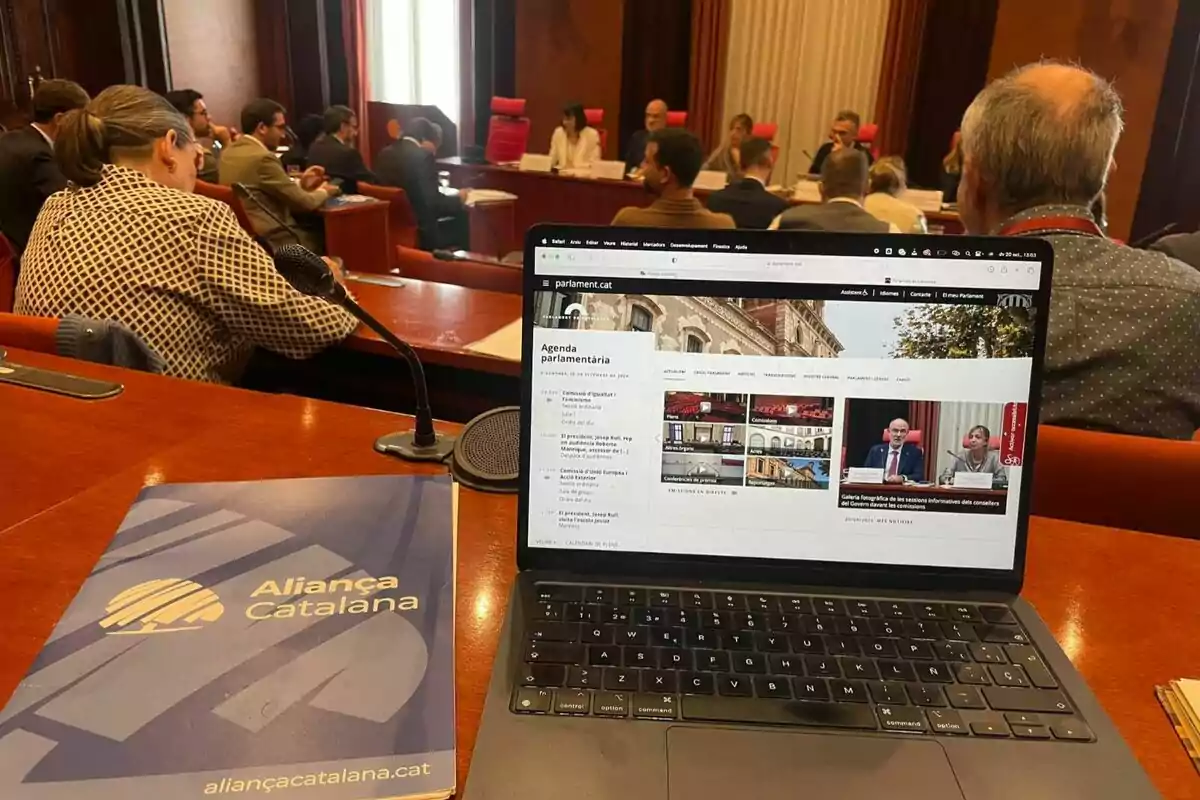 Una portàtil oberta mostrant una pàgina web d'un parlament, amb una carpeta d'Aliança Catalana al costat, en una sala de reunions amb diverses persones assegudes al voltant d'una taula.