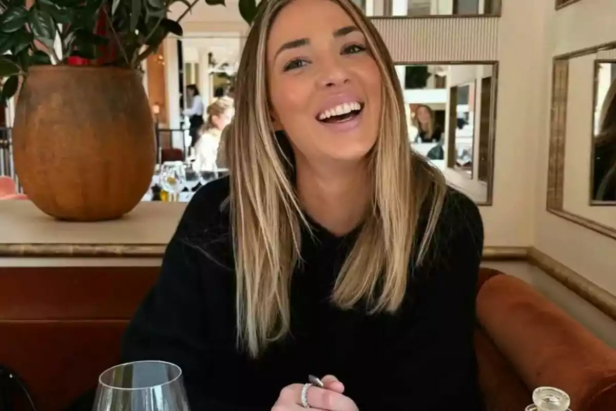 Una mujer sonriente con cabello rubio y suelto está sentada en un restaurante, con una planta en el fondo y copas sobre la mesa.