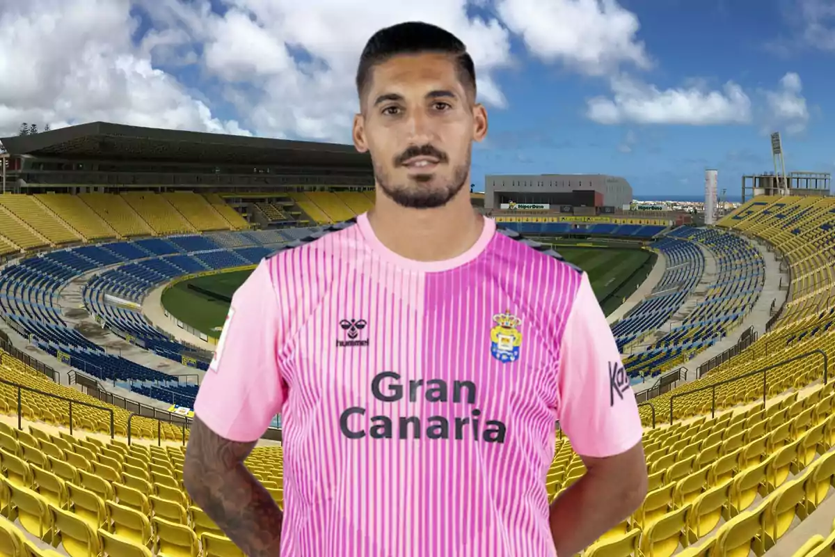 Un jugador de fútbol con una camiseta rosa de la UD Las Palmas posando en un estadio vacío con asientos amarillos y azules.