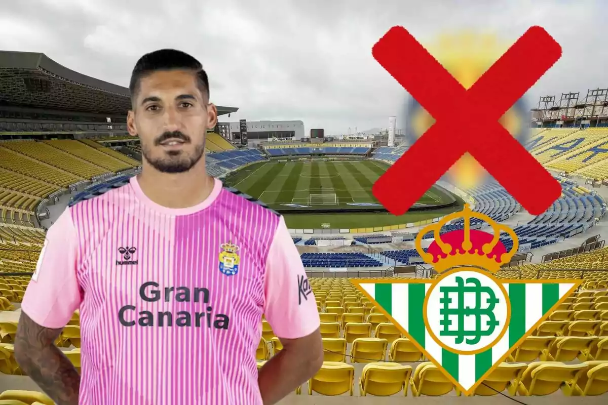 Un jugador de futbol amb una samarreta rosa de Gran Canària està dreta en un estadi buit amb seients grocs i blaus, amb un gran símbol de X vermella i l'escut del Real Betis a la cantonada inferior dreta.