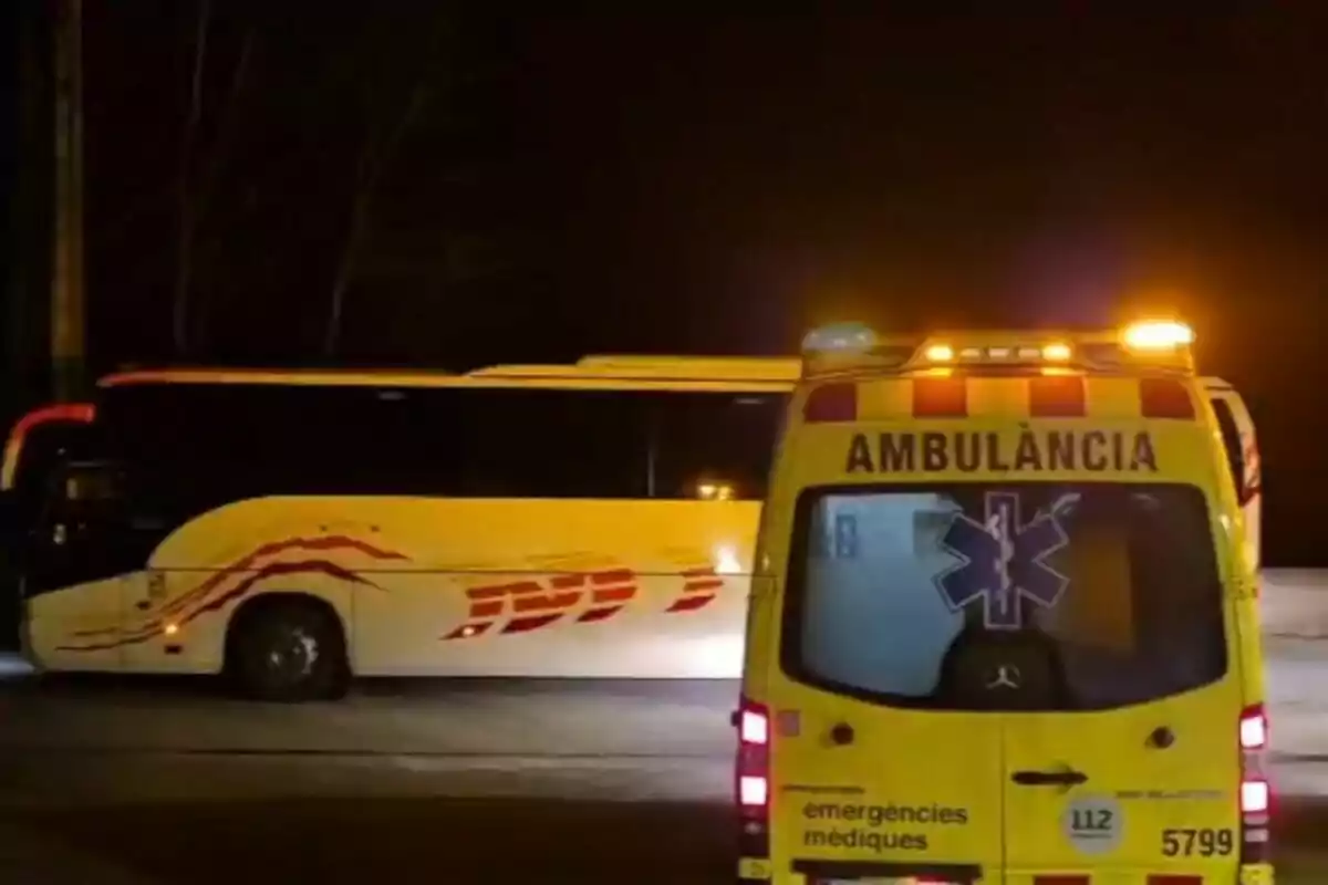 Una ambulància groga amb llums encesos està estacionada darrere d'un autobús blanc amb detalls vermells en una carretera fosca.