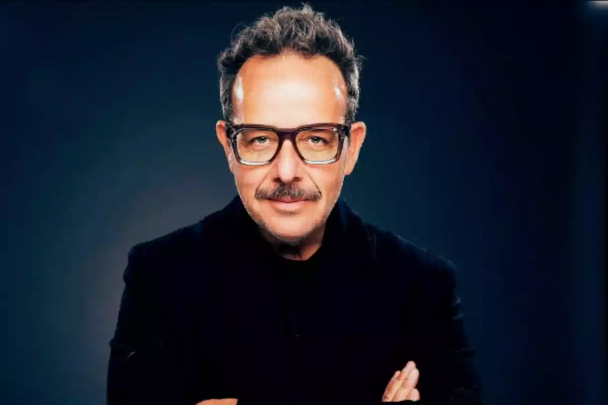 Un hombre con gafas y bigote, vestido de negro, posando con los brazos cruzados frente a un fondo oscuro.