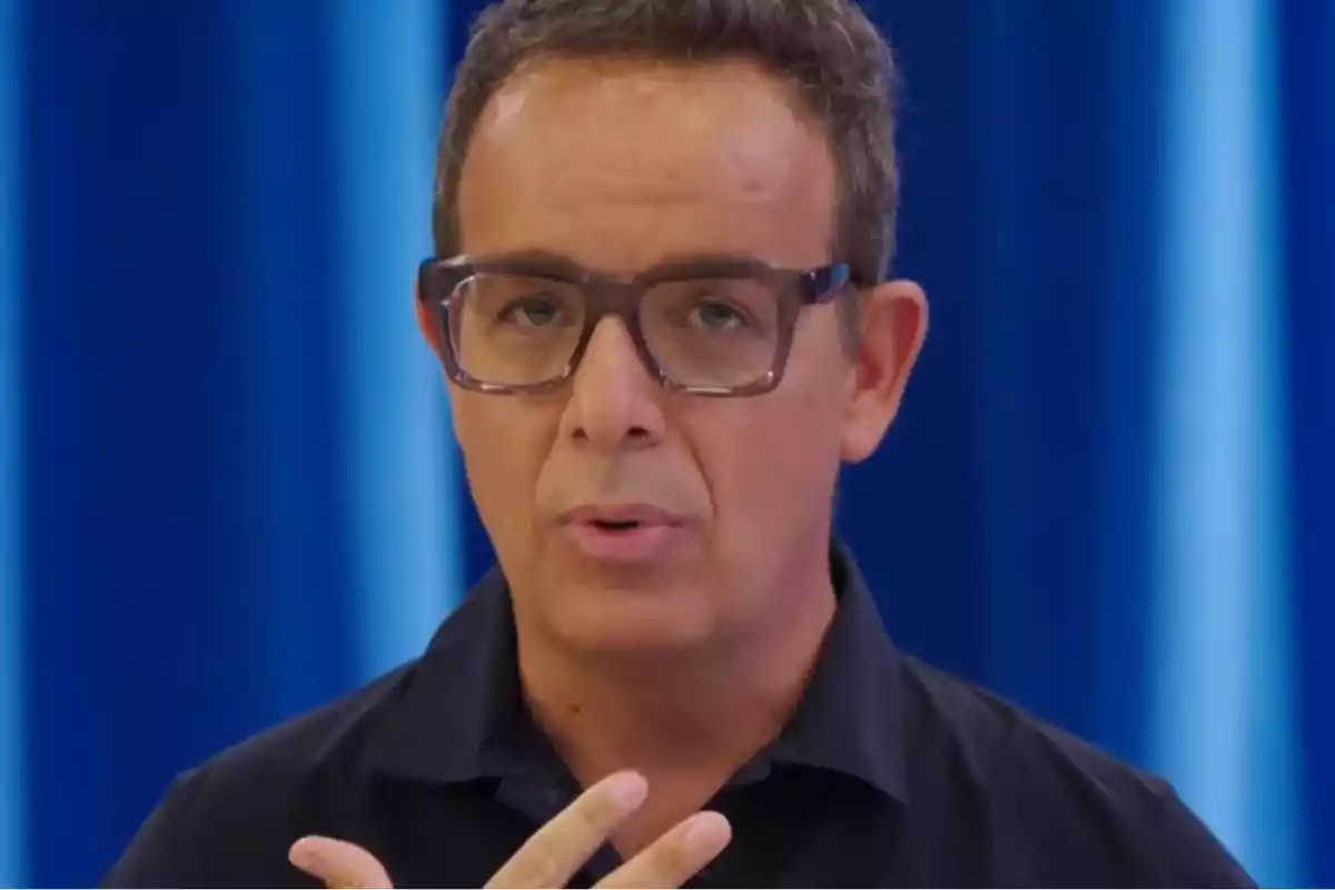 Hombre con gafas y camisa oscura hablando frente a un fondo azul.