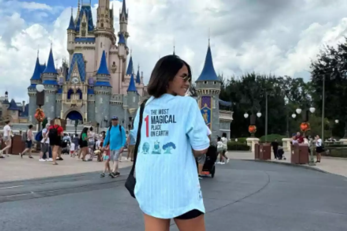 Una mujer con gafas de sol y camiseta azul claro está de pie frente a un castillo en un parque temático, rodeada de visitantes y un cielo nublado.