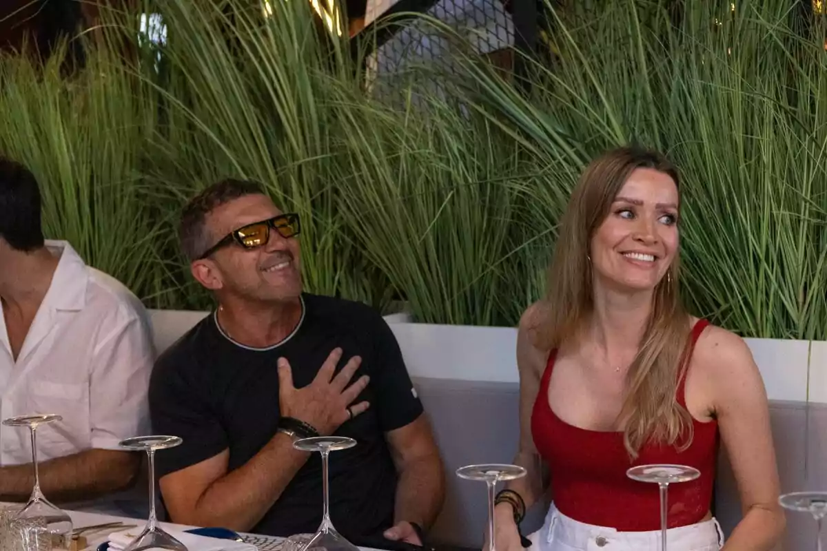 Antonio Banderas y Nicole Kimpel sonrientes sentados en una mesa con copas de vino vacías, rodeadas de plantas verdes. Antonio Banderas y Nicole Kimpel sonrientes sentados en una mesa con copas de vino vacías, rodeadas de plantas verdes.
