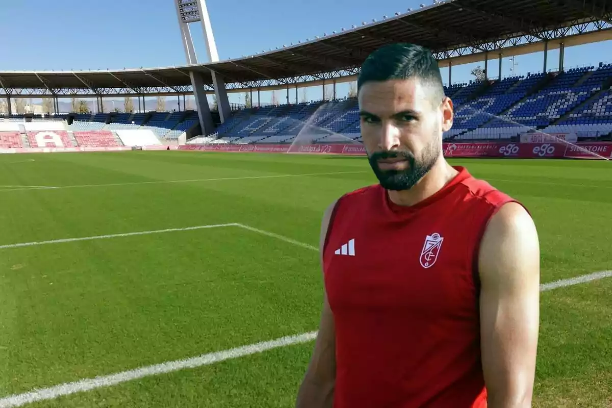 Un jugador de fútbol con camiseta roja posando en un estadio vacío con césped verde y gradas en el fondo.