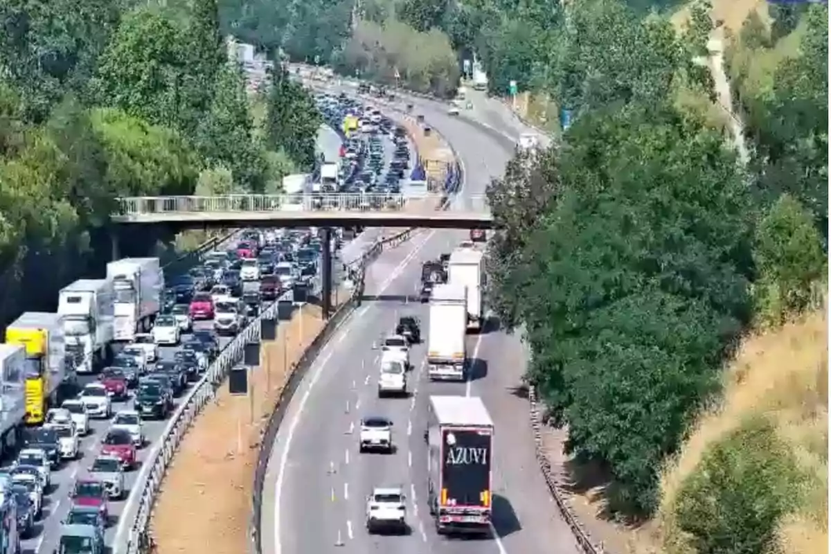 Carretera amb trànsit dens en un sentit i circulació fluida en l’altre, envoltada d’arbres i amb una passarel·la per a vianants creuant per sobre dels carrils.